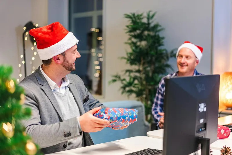 Two men in Santa hats exchanging gifts in a decorated office setting.